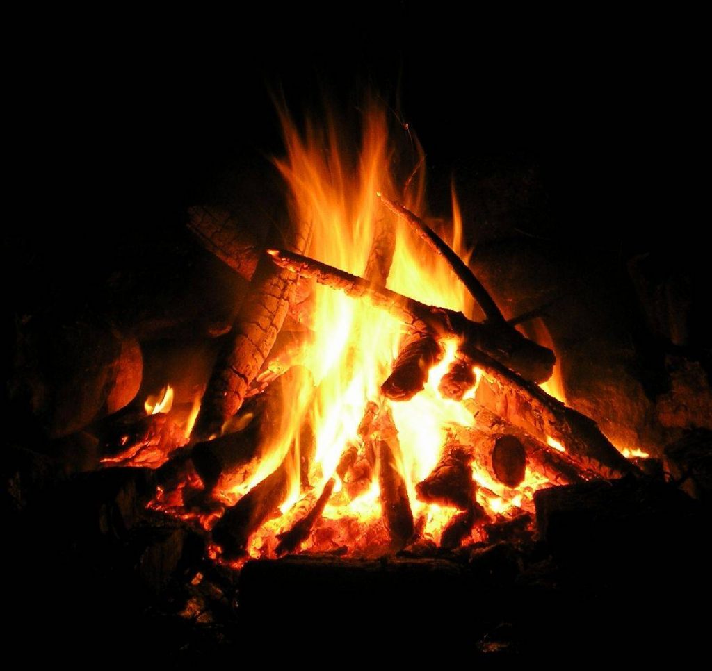 A campfire burns brightly in the dark, with orange and yellow flames rising from glowing logs and embers, surrounded by darkness.
