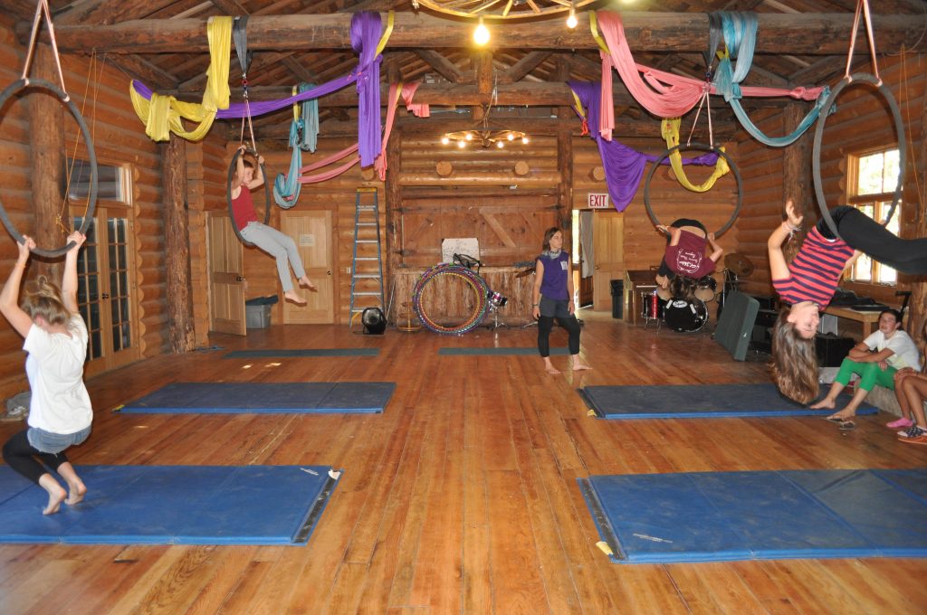 Children practice aerial hoops and acrobatics on mats inside a rustic wooden cabin with colorful fabric draped overhead. Some kids swing on hoops, while others watch or help each other near the back of the room.
