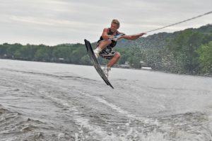 A person wearing a life jacket and shorts is wakeboarding, catching air above the water while holding onto a tow rope, with trees and a cloudy sky in the background.