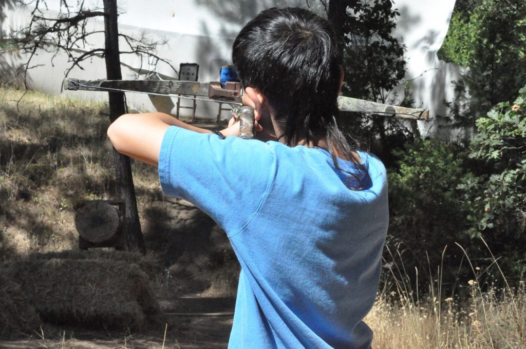 A person with dark hair wearing a blue shirt aims a crossbow outdoors, targeting a distant bullseye in a wooded area—a perfect scene for family camp activities among hay bales and dappled sunlight.