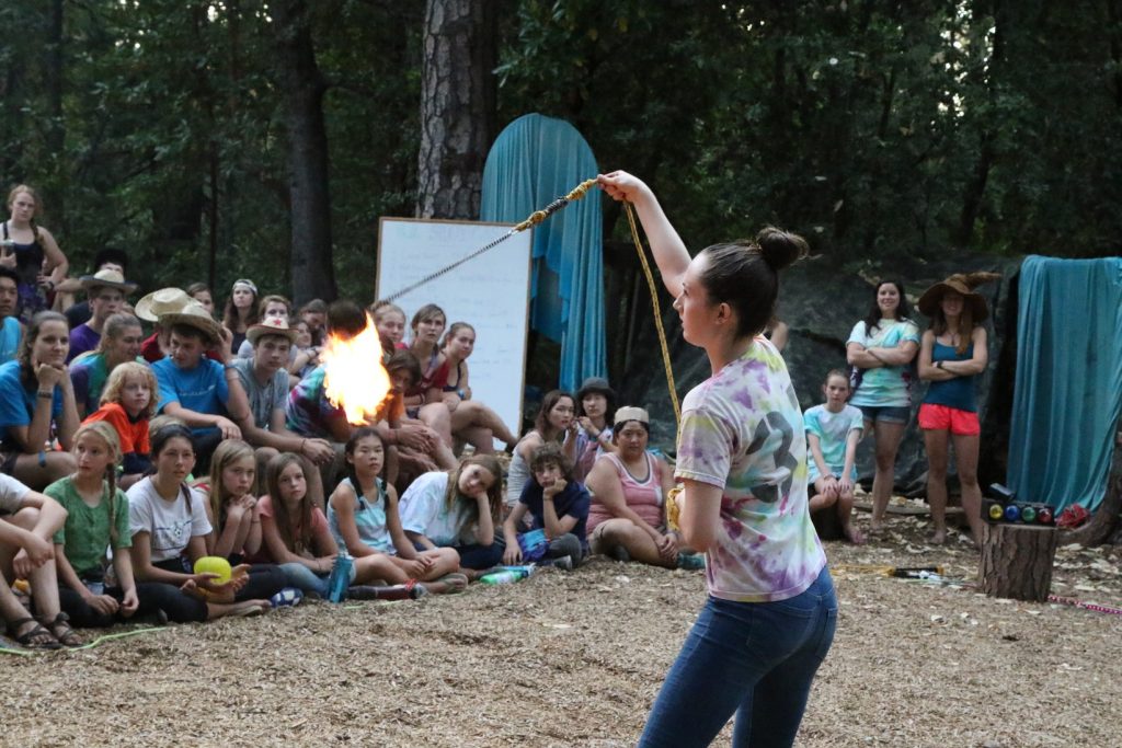 A woman in a tie-dye shirt swings a flaming rope before a seated audience of children and adults outdoors, surrounded by trees and blue fabric decorations.