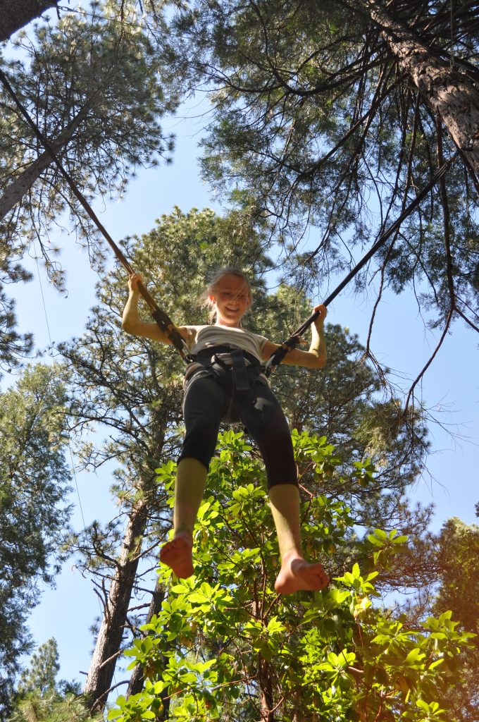 A smiling child is suspended on a harness and ropes among tall pine trees, seen from below—enjoying family camp activities. Sunlight filters through the leaves, highlighting the child's bare feet and joyful expression.