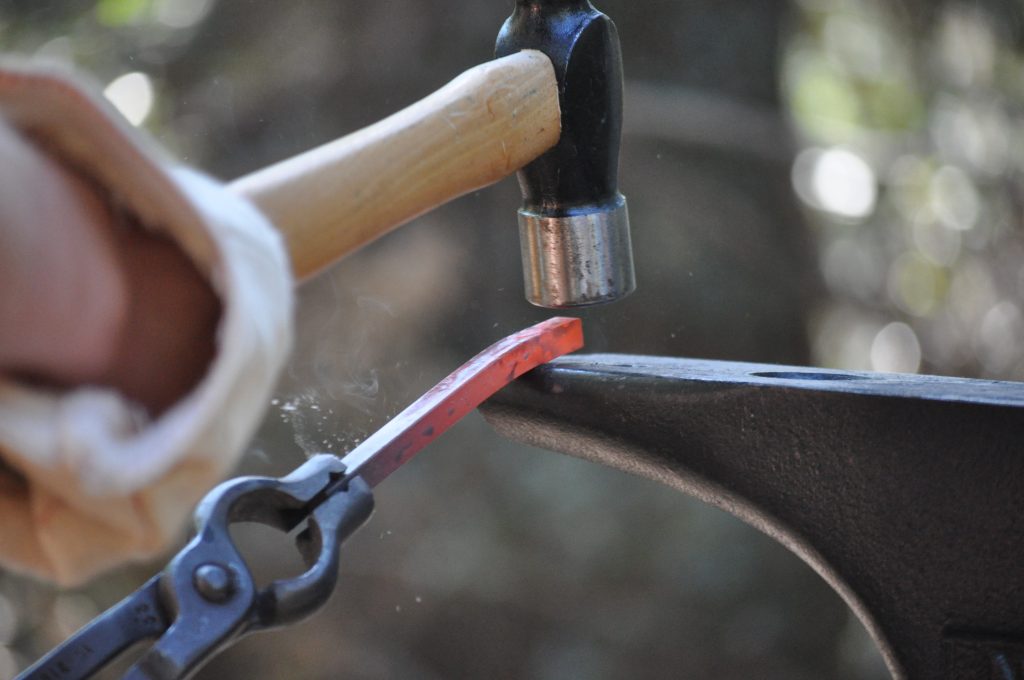 A close-up of a person using tongs to hold a hot metal rod on an anvil while striking it with a hammer, demonstrating blacksmithing—a popular choice for hands-on family camp activities—with steam or smoke rising from the heated metal.