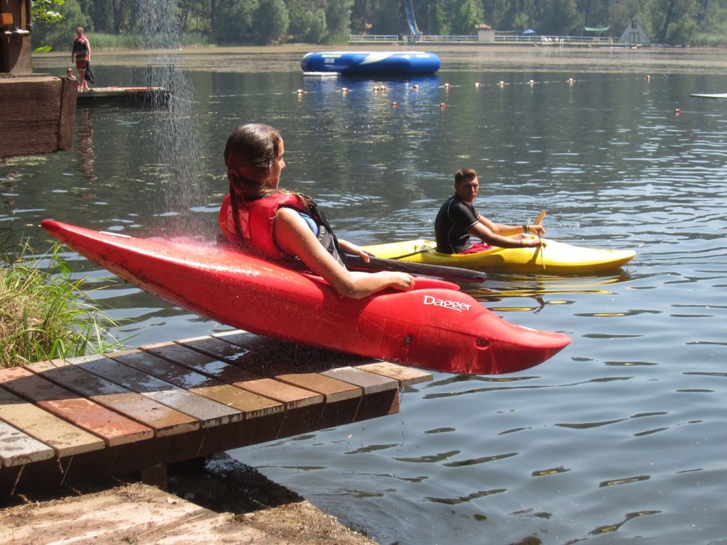 A person in a red kayak slides off a dock into a lake during family camp activities, wearing a life vest, while another kayaker floats nearby. Trees and inflatable water equipment can be seen in the background.