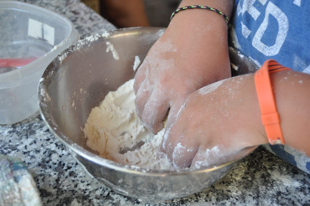 A person mixes flour with their hands in a metal bowl on a countertop, wearing a blue shirt and colorful wristbands—perfect for fun family camp activities in the kitchen.