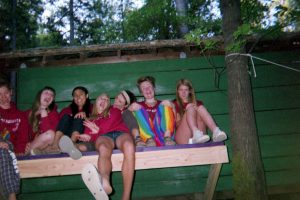 A group of seven teenagers sit closely together on a wooden platform attached to a green wall outdoors, smiling and laughing. Trees and leaves surround them in the background.