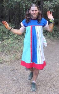 A person with long hair and a beard is wearing a colorful dress and green sneakers, standing on a dirt path in a wooded area, raising both hands playfully with orange stains on their fingers and arms.