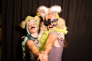 Two people in costume pose together against a black curtain. One wears a cowboy hat, fake mustache, and scarf, while the other wears a bunny hat, sunglasses, blonde wig, and feather boa. Both have their arms outstretched.
