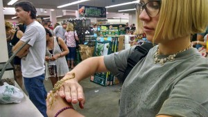 A person with blonde hair and glasses holds a large insect, possibly a mantis, on their hand at an indoor event with people and reptile supplies in the background.