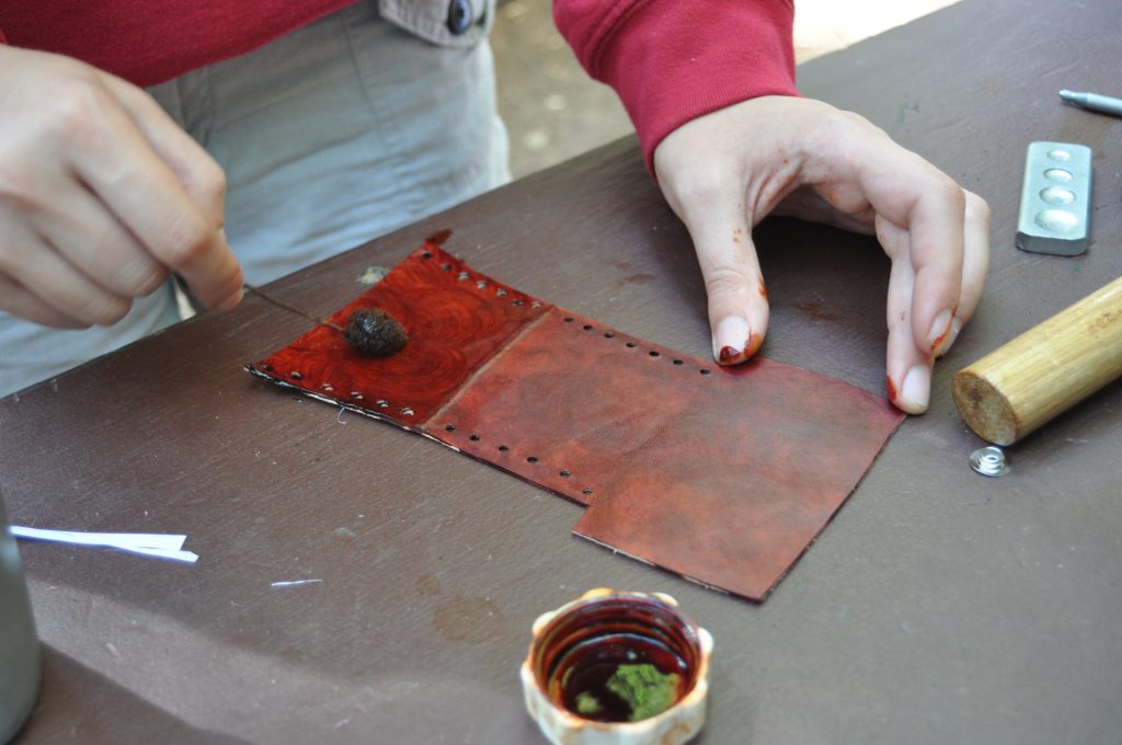 A person applies dark stain to a piece of brown leather with a cloth, preparing it for leathercraft. Holes are punched along one edge, and various tools sit on the table nearby.