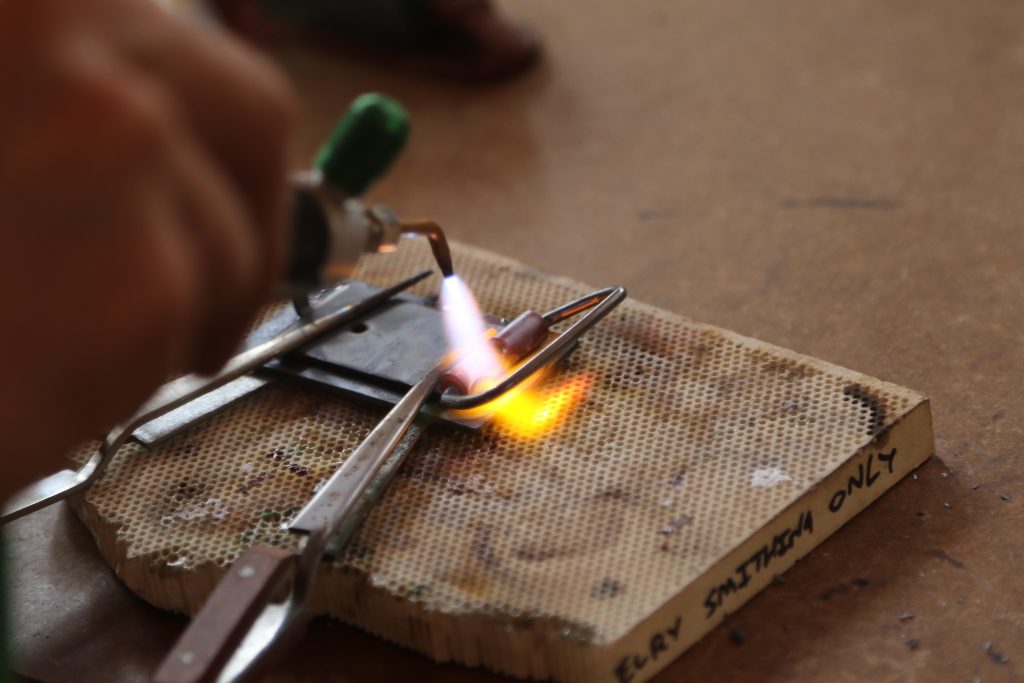 A person uses a small blowtorch to heat a metal object held by pliers on a ceramic work surface labeled “FLUX SOLDERING ONLY”—a hands-on project often featured in family camp activities. The flame produces a bright orange-yellow glow on the metal.