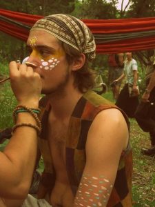 A young person wearing a colorful, sleeveless vest and headscarf sits outdoors while someone paints white and yellow patterns on their face and arms. There are trees and people in the background, and a red hammock is hanging behind them.