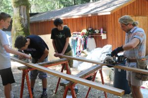 Four people work together outdoors, cutting long wooden boards on sawhorses—a scene reminiscent of past cabin activities. Clothing hangs on a rack by a wooden shed, while surrounding trees provide shade for the busy workspace.