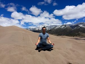 A person sits cross-legged and smiling on sandy dunes under a blue sky with scattered clouds, with snow-capped mountains and trees in the background.