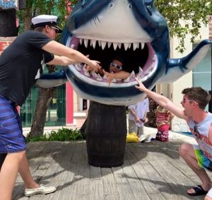 Three people pose playfully with a large shark statue. One person sits inside the shark’s mouth, while the other two stand outside, reaching toward the person in the mouth, as if trying to rescue them.