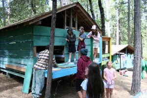 A group of children stands and talks outside a rustic, green wooden cabin in a forest, reminiscing about past cabin activities. Some are on the porch while others are on the ground. Trees surround them, and colorful towels hang nearby.