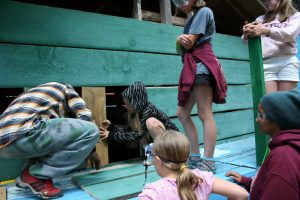 A group of children, some kneeling and some standing, work together to build or repair a wooden structure—much like in past cabin activities. They wear casual clothes and hoodies or sweatshirts, focusing intently on the task.