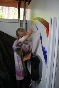 An elderly person painting a colorful mural with a yellow star and vibrant streaks on a white wall, holding a paint palette and wearing a paint-splattered shirt—reminiscent of past cabin activities.