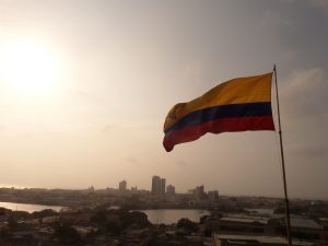 A Colombian flag waves on a pole against a hazy sky, with a city skyline and river visible in the background at sunset.