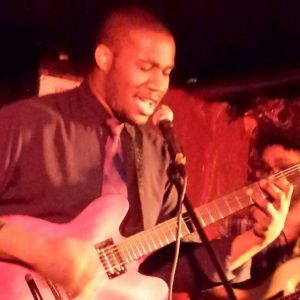A man passionately sings into a microphone while playing an electric guitar on stage under warm, dim lighting.
