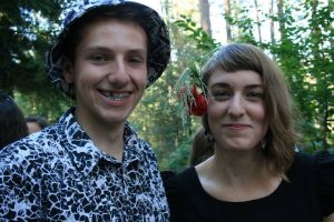 Two people smiling outdoors with trees in the background. The person on the left wears a black-and-white patterned shirt and bucket hat. The person on the right has short hair and a red flower hair accessory.