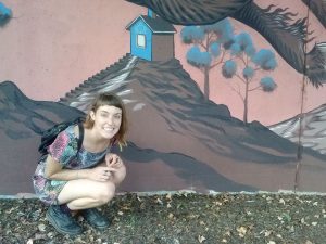 A smiling person with short brown hair crouches in front of a mural showing a blue house on a hill with trees. The person wears a patterned dress, black shoes, and a backpack, and there are leaves on the ground.