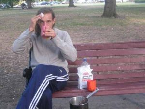 A man sits on a park bench, drinking from a cup and wiping his forehead with a napkin. A plastic bag of sugar, an orange mug, and a pot rest on the bench beside him. Trees and grass are visible in the background.