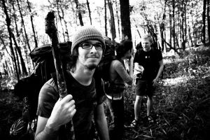 A young man with glasses and a beanie holds a hiking stick and smiles at the camera in a forest. In the background, three people stand and talk among the trees. The photo is in black and white.