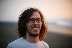 A young man with long brown hair, glasses, and a beard smiles while standing outdoors, with a blurred background of sand and sky suggesting a beach at sunset.