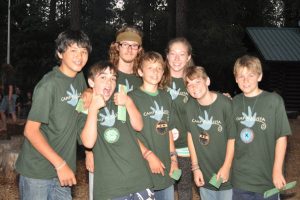 A group of smiling kids and one adult, all wearing matching green Camp Augusta shirts, pose together outdoors with trees and a cabin in the background. Some are holding cards and wearing name tags.