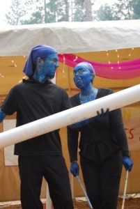 Two people dressed in black with blue-painted skin and faces, wearing blue head coverings, stand outdoors under a tent. One holds a large white tube. Pink fabric and string lights hang behind them.
