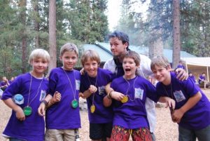 Six children and an adult, all wearing purple t-shirts and handmade name tags, pose happily together in a forested camp area with tents and people in the background.