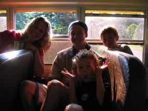 Four people, two adults and two children, are sitting and smiling inside a school bus with sunlight streaming through the windows. One child in front is making a playful gesture with their hands.