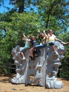 Four people sit and pose with arms raised on top of a gray playground climbing wall, surrounded by trees and bright sunlight in an outdoor park setting.