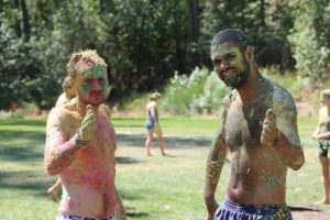 Two people outdoors, smiling and covered in colorful paint, give a thumbs-up gesture. They stand on grass with trees and blurred figures in the background, suggesting a fun outdoor event or activity.
