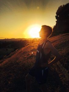 A person in athletic clothing stands on a hillside at sunset, posing with hands on hips and head tilted back, with the sun setting behind them and trees in the background.