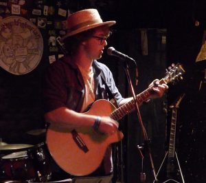 A person wearing a straw hat and glasses plays an acoustic guitar and sings into a microphone on a dimly lit stage with a drum set and an electric guitar in the background.