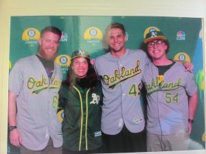 Four smiling people wearing Oakland Athletics baseball jerseys and caps pose together in front of a green backdrop with Oakland Athletics and TV logos.
