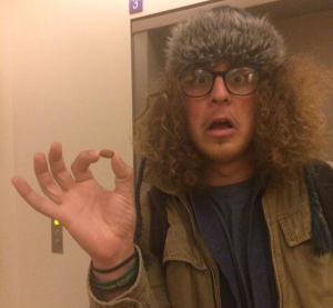 A person with curly hair, glasses, and a furry hat looks surprised while holding a small round object between their fingers in front of an elevator.