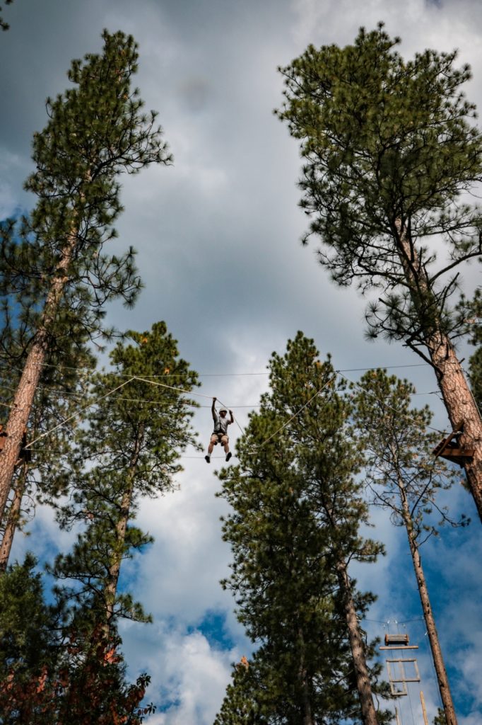 A person rides a zipline high among tall pine trees under a partly cloudy sky, surrounded by green foliage and wooden platforms attached to the trees.