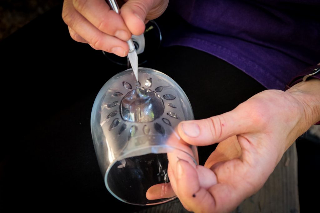 A person uses a sharp tool to carve or etch decorative patterns onto the base of a clear glass, holding the glass steady with one hand.