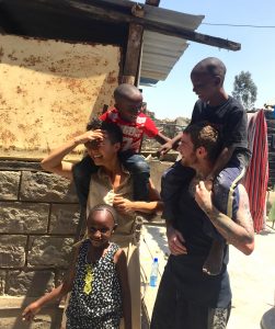A smiling woman and man each carry a child on their shoulders, while a young girl stands in front of them. They are outdoors by a stone building, sharing a joyful moment together in the sunlight.