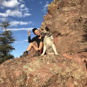 A woman in hiking gear sits on a rocky outcrop next to a happy dog, both smiling under a bright blue sky with scattered clouds. A pine tree is visible to the left of the large reddish rocks.