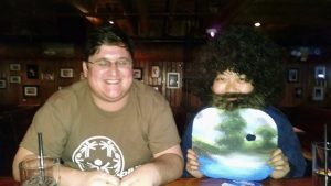 Two people sit at a wooden table in a dimly lit bar. One is smiling and wearing glasses with a brown T-shirt; the other is in a Bob Ross costume, holding a palette and wearing a curly wig and fake beard.