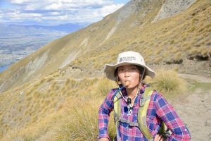 A hiker wearing a sun hat, plaid shirt, and backpack drinks from a hydration tube while standing on a grassy mountain trail with scenic hills and a valley in the background under a partly cloudy sky.