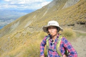 A person wearing a hat, plaid shirt, and backpack drinks from a hydration tube while hiking on a sunlit mountain trail, with grassy slopes and a valley visible in the background.