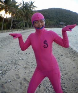 A person in a full-body pink suit with a hood and gloves stands on a beach, making a playful pose with their hands up and palms facing out. Palm trees and a hillside are visible in the background.