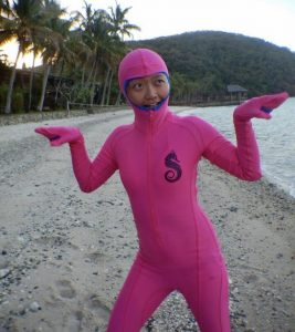 A person in a full-body pink suit and hood stands on a sandy beach with their arms raised and palms up, with palm trees and a hill in the background.