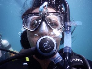 A close-up view of a person underwater wearing scuba diving gear, including a mask and regulator, with air bubbles rising around their face and another diver visible in the background.