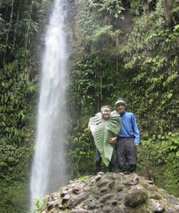 Two people stand on rocks in front of a tall waterfall surrounded by lush greenery. One person is holding a large leaf, partially covering themselves, while the other stands beside them, both smiling at the camera.